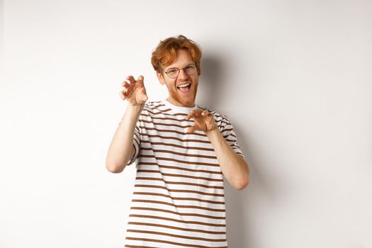 Funny young man with red hair and beard showing claws gesture and roaring, having fun, standing happy over white background.