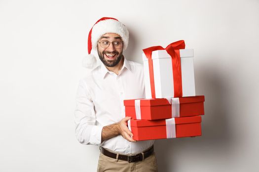 Merry christmas, holidays concept. Excited man celebrating xmas, wearing santa hat and holding gifts, standing over white background.