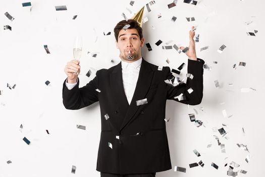 Handsome party guy in black suit having fun, celebrating new year, blowing whistle and drinking champagne while confetti falling, wishing happy holidays, standing against white background.