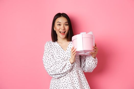 Valentines day. Happy asian woman receiving gift in cute box, smiling excited and thankful, standing in dress against pink background.