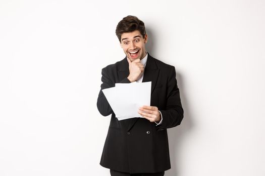 Portrait of handsome businessman looking excited at documents, working, standing against white background in black suit.