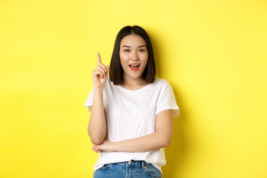 Beauty and fashion concept. Excited asian girl raising finger in eureka gesture, pitching an idea and smiling, standing over yellow background.
