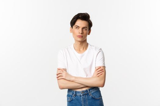 Thoughtful gay man in crop top looking at upper left corner, thinking with arms crossed on chest, standing against white background.