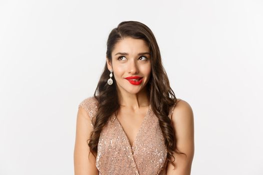Close-up of elegant and beautiful woman in dress and red lipstick thinking, looking at upper left corner and biting lip from temptation, standing over white background.