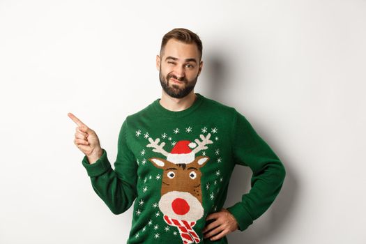 New year celebration and winter holidays concept. Skeptical guy in christmas sweater looking displeased, pointing finger at upper left corner product, dislike it, white background.