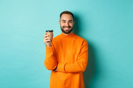 Handsome smiling man taking break, drinking coffee from takeaway, standing over blue background.