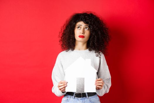 Real estate. Sad woman dreaming of buying apartment, holding paper house cutout and looking up distressed, standing on red background.