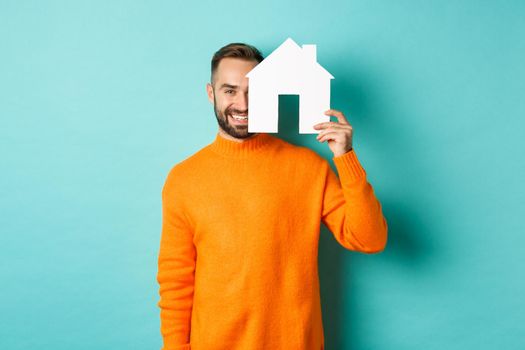Real estate concept. Happy young man searching for home rent, holding house paper maket and smiling, standing over blue background.