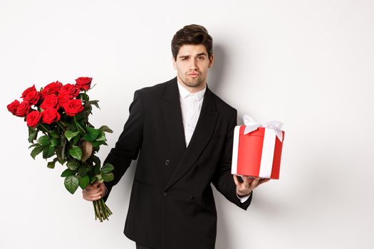 Concept of holidays, relationship and celebration. Handsome and confident man in black suit, going on a date, holding bouquet of roses and present, standing against white background.