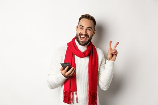 Happy bearded man using smartphone, posing for photo with peace sign, standing in winter sweater and red scarf, white background.