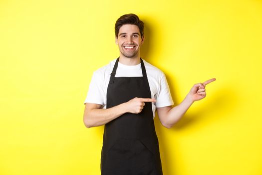 Friendly waiter pointing fingers right, showing your logo or promo offer, wearing black apron uniform, standing over yellow background,