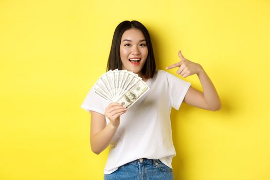 Smiling asian woman pointing hands at money, showing dollars, standing over yellow background.
