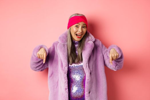 Fashion and shopping concept. Excited asian elderly woman checking out special deal, pointing fingers down and smiling happy at camera, showing advertisement, pink background.