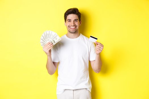 Young man withdraw money from credit card, smiling pleased, standing over yellow background.