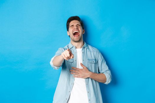 Adult man in casual outfit laughing out loud and pointing at you, looking at something funny, chuckling while standing against blue background.