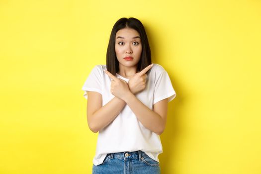 Indecisive asian girl need help with choice, pointing fingers sideways and looking confused, standing over yellow background.