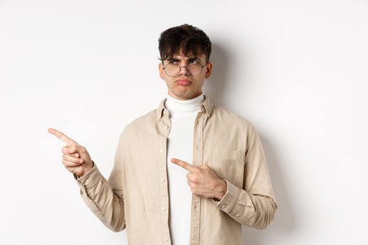 Disappointed guy in glasses looking and pointing left, sulking displeased, complaining on bad offer, standing upset on white background.