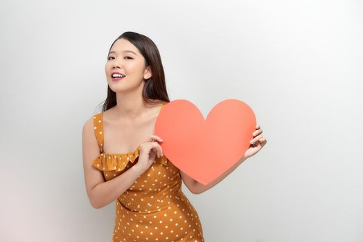 smiling pretty woman looking upwards, holding large red heart to chest daydreaming of men in love,