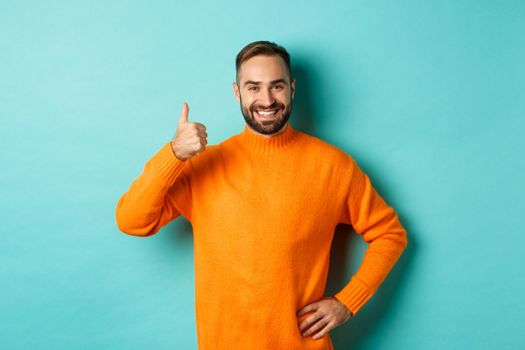 Well done. Handsome bearded man showing thumb up, praising good work, recommend excellent product, like and approve, smiling satisfied, standing over light blue background.