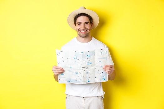 Travelling, vacation and tourism concept. Smiling young man going on trip, holding road map and smiling, standing over yellow background.