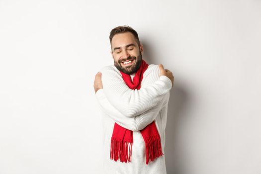 Happy bearded man enjoying comfortable winter sweater, smiling with warmth, hugging himself, standing over white background.