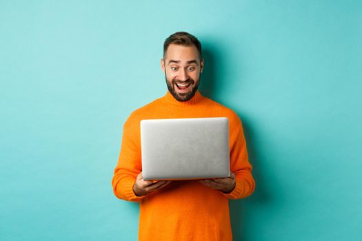 Excited man in orange sweater working on laptop, looking at computer screen amazed, standing over light blue background.
