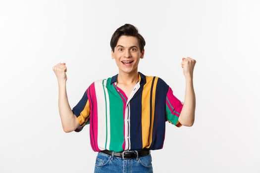 Happy young man rejoicing, clench fists and looking with satisfaction, achieve goal or win prize, standing over white background.