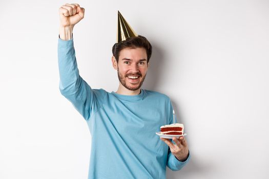 Celebration and holidays concept. Happy man celebrating birthday in party hat, holding bday cake and raising hand up in triumph, standing over white background.