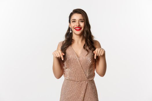 Happy beautiful woman with red lips, smiling while showing promo, wearing elegant dress for christmas party, pointing fingers down, standing over white background.