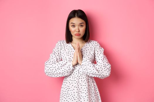 Cute asian girl asking for help, begging for favour and looking innocent at camera, pleading against pink background.