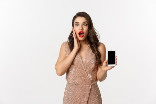 Online shopping. Elegant woman in luxury dress showing smartphone screen, looking amazed at camera, white background.