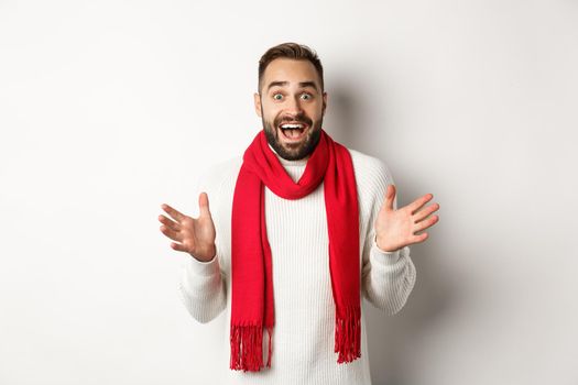 Christmas holidays and celebration concept. Bearded guy looking surprised at new year promo offers, gasping amazed, wearing red scarf and sweater, white background.