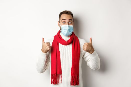 Covid-19, lockdown and quarantine concept. Surprised man in face mask showing thumbs-up, standing in sweater and red scarf, white background.