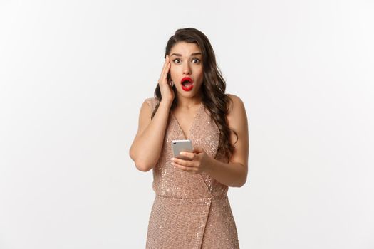 Holidays, online shopping concept. Surprised woman using mobile phone and looking amazed, wearing party dress, standing over white background.