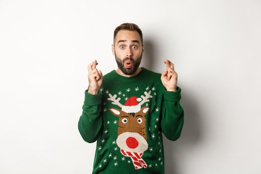 Christmas, holidays and celebration. Excited guy making wish, keep fingers crossed for good luck and smiling, standing over white background.