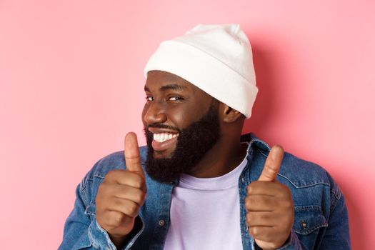 Close-up of happy Black bearded guy in beanie showing support, agree or approve something, giggle devious and showing thumbs-up, standing over pink background.