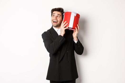 Concept of christmas holidays, celebration and lifestyle. Image of excited man enjoying new year, shaking gift box to guess what inside, standing against white background.