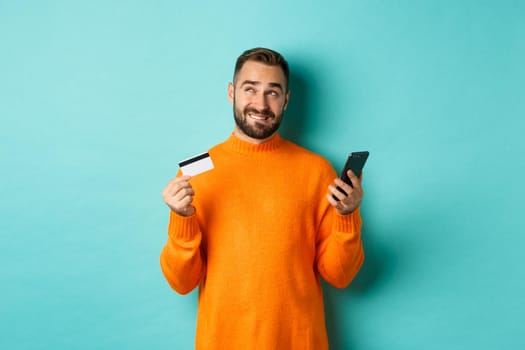 Online shopping. Thoughtful man holding credit card and mobile phone, thinking of purchase, standing over light blue background.