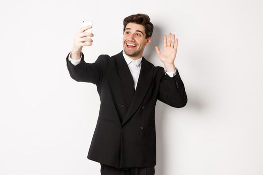 Image of handsome man in suit, having video call and waving hand at smartphone camera, recording video, greeting someone, standing against white background.