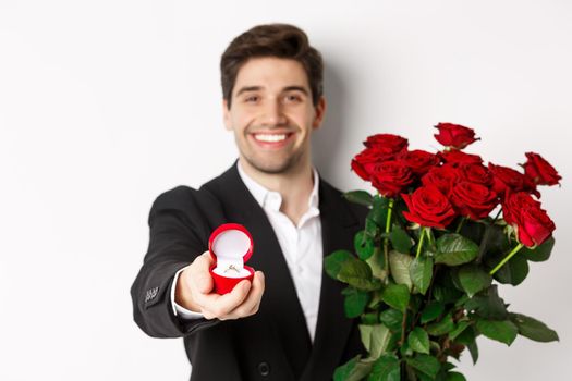 Close-up of attractive man in suit, holding bouquet of roses giving an engagement ring, proposing to girlfriend, standing against white background.