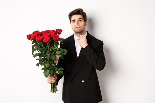Image of elegant and sassy man in black suit, looking confident and holding bouquet of red roses, going on a romantic date, standing against white background.