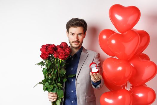 Handsome man in suit giving an engagement ring and bouquet of red roses, marry me on Valentines day, standing with heart balloons on white background.
