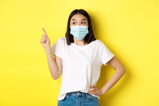 Covid, health care and pandemic concept. Asian woman in medical mask and white t-shirt pointing finger at upper left corner logo, showing promotion, yellow background.