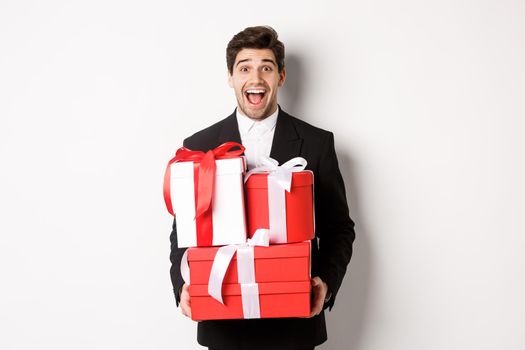 Concept of christmas holidays, celebration and lifestyle. Image of handsome amazed guy in suit, holding new year presents and smiling, standing against white background.