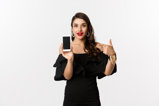 Online shopping concept. Fashionable woman in black dress pointing finger at smartphone screen, showing application, standing over white background.