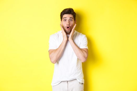 Image of handsome man looking surprised, gasping and saying wow, looking at promo offer amazed, standing over yellow background.