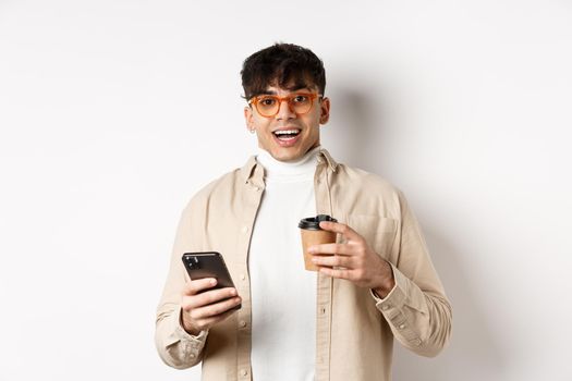 Cheerful smiling young man in glasses drinking coffee and holding smartphone, looking excited at camera, standing on white background.