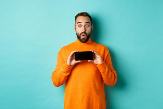 Impressed man showing smartphone screen, stare at camera amazed, demonstrate display, standing over light blue background.
