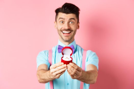 Valentines day. Romantic young man in bow-tie stretch hands with engagement ring and smiling, asking to marry him, making proposal, standing over pink background.