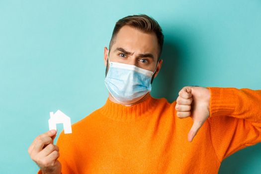 Real estate and coronavirus pandemic concept. Close-up of disappointed man in face mask, showing small paper house and thumb-down, turquoise background.